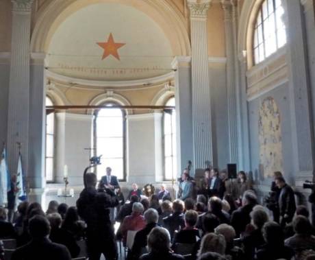Pohled na průčelí kostela s mluvčím vepředu a publikem sedícím na židlích